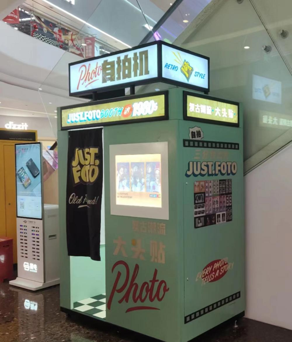 图片展示是大头贴机器的实物展示。机器主体为绿色,上方有“Photo自拍机”字样,侧面写有“JUST FOTO”等标识。机器前方设有拍照窗口,窗口内有照片展示。机器底部有“大头贴”字样,旁边还有“EVERY PHOTO TELLS A STORY”等装饰文字。该图片与上下文紧密相关,直观呈现了大头贴机器的外观样式,为介绍其痛点及后续改造提供视觉参考。