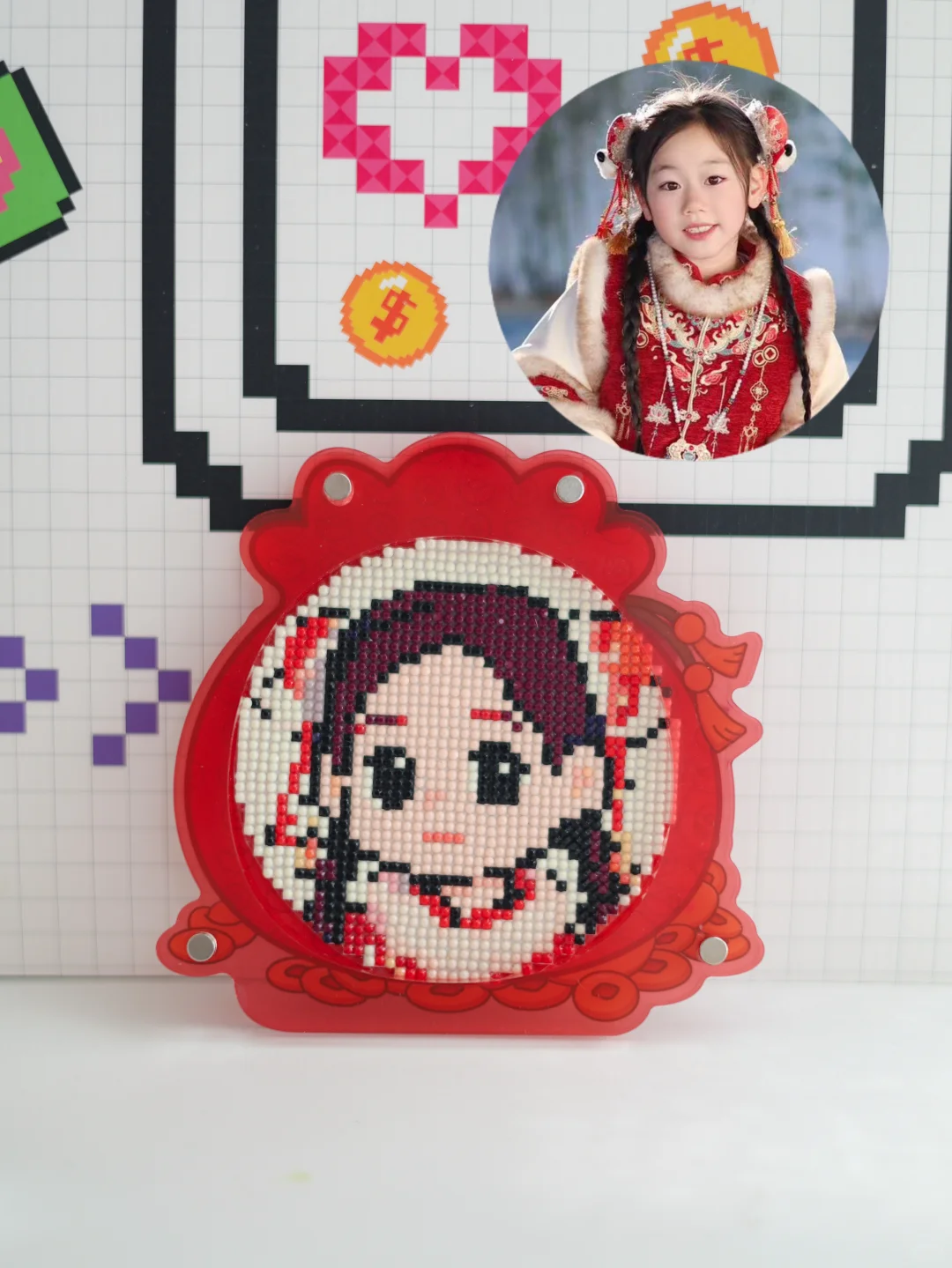 图片展示了一幅钻石画作品,画面中是一个小女孩的卡通形象,背景为红色。作品被放置在一个红色的圆形底座上,底座边缘有红色的装饰。背景墙上有网格图案,左侧有紫色、绿色的图案,右侧有粉色、黄色、橙色的图案,还有一张小女孩的真人照片。该图片与文档中介绍钻石画的内容相关,直观呈现了钻石画的成品效果。