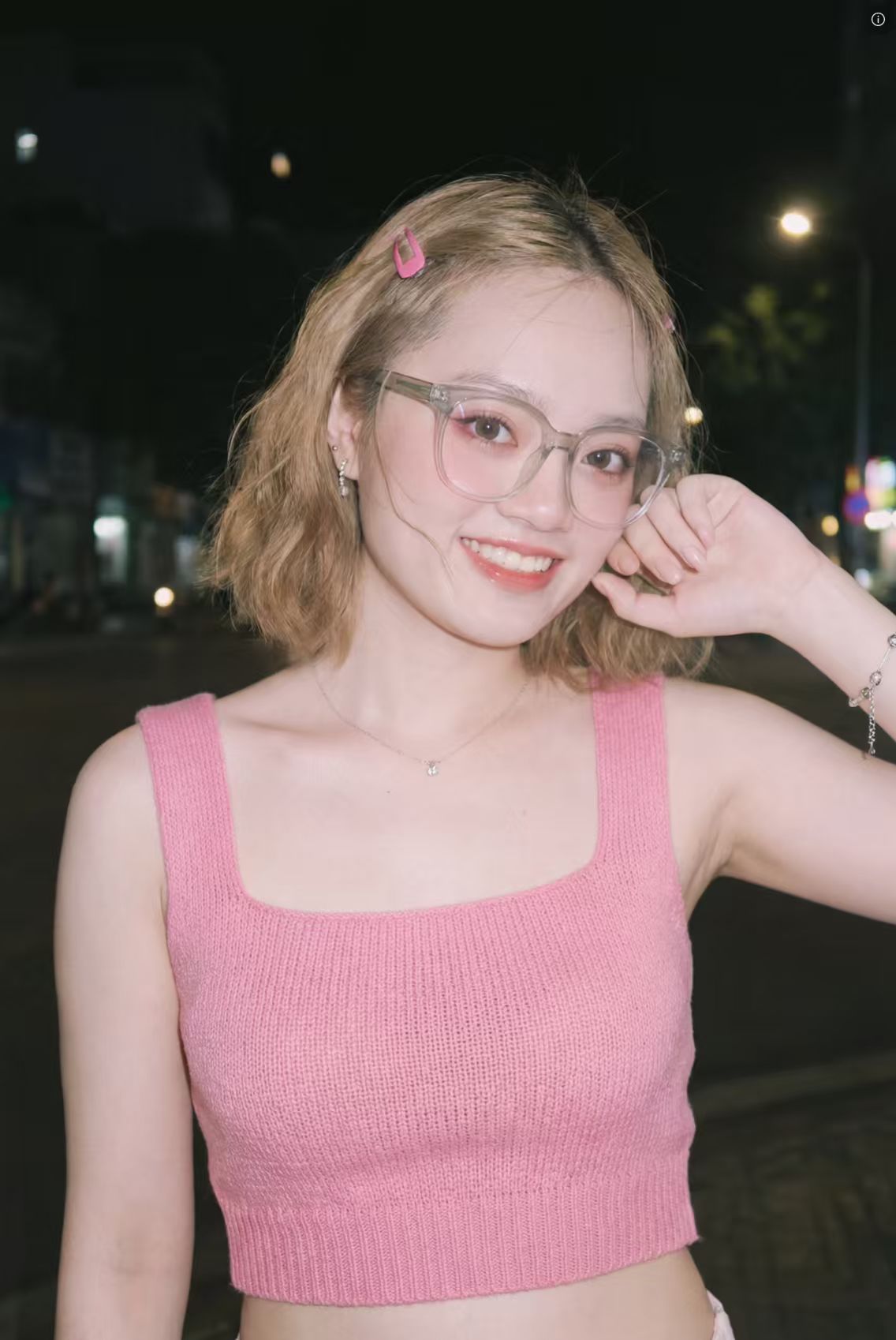 图片展示了一位年轻女性,她留着浅棕色短发,头发一侧别着粉色发卡,戴着透明边框眼镜,身穿粉色针织吊带背心,面带微笑,姿态轻松自然。背景是夜晚的街道,有路灯和模糊的街景。结合上下文可知,这是一张通过相关技术生成和优化后的照片效果示例,展示了仅凭一张拍完的图片,就能产出类似影楼效果照片的“影楼级”生产力下放成果。