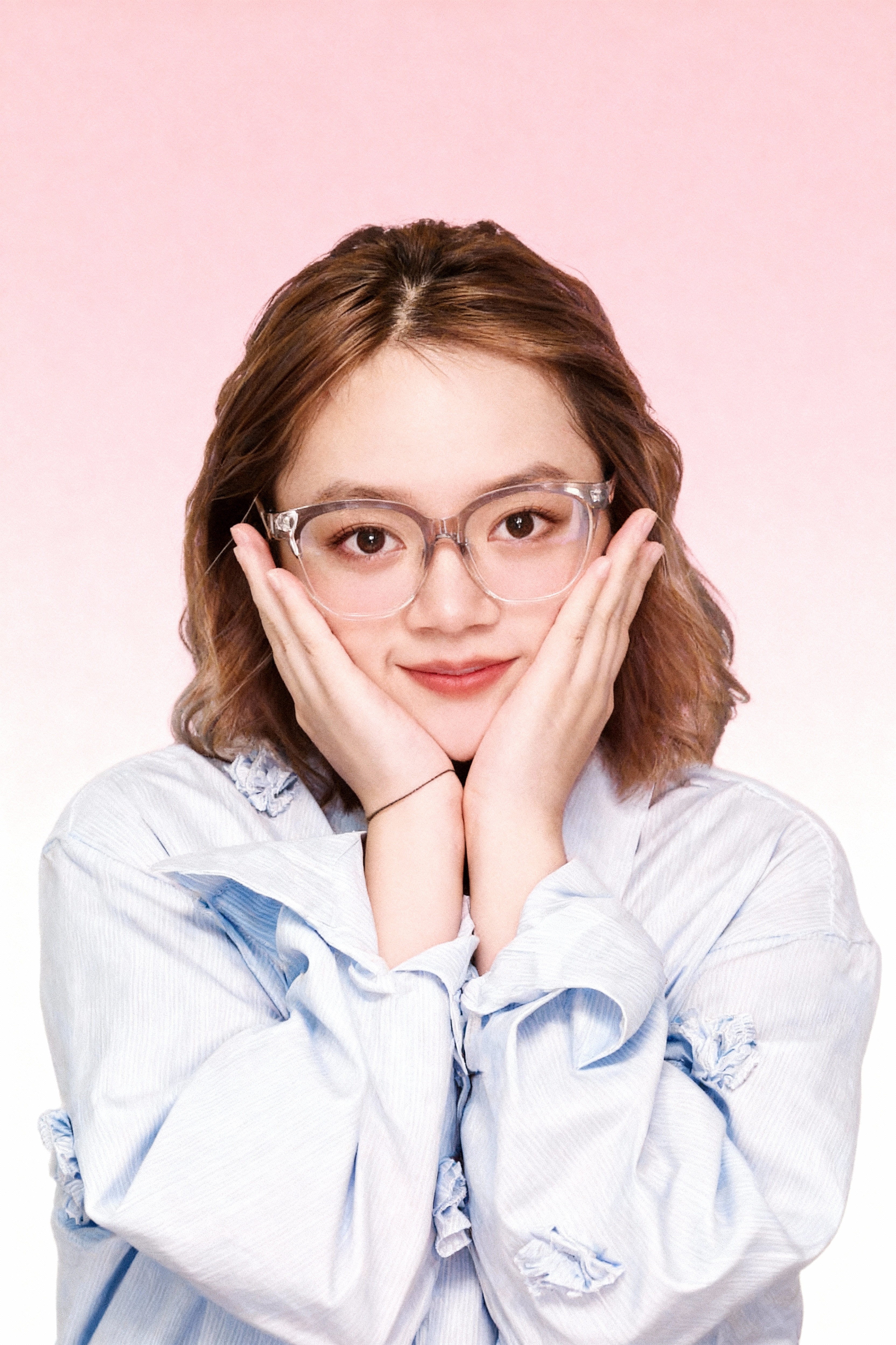 图片展示了一位年轻女性,她戴着透明框架眼镜,双手托腮,面带微笑,表情自然。她穿着浅蓝色带有白色花纹的衬衫,背景为渐变色,从粉色过渡到白色。该图片是文档中“大头贴机器——存量机器焕发新生”案例中生成图对比的一部分,用于展示通过AI技术优化后的效果,与上下文提到的“仅凭一张拍完的图片,就可以生成和优化出数十套原本在摄影馆要花大几百才能产出的照片效果”相呼应。