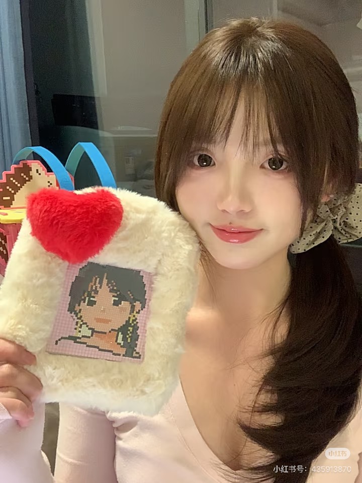图片展示了一位年轻女性,她有着棕色长发,戴着白色发带,穿着粉色上衣。她左手拿着一个毛绒材质的包,包上有一个像素风格的卡通人物图案,图案中人物有黑色头发,背景是粉色和白色。图片右下角有“小红书号:429938290”字样。该图片与文档中“蒂彩的定制业务已经能在小红书上通过‘Doo crafts旗舰店’直接体验”的内容相关,展示了在小红书上体验定制业务的场景。