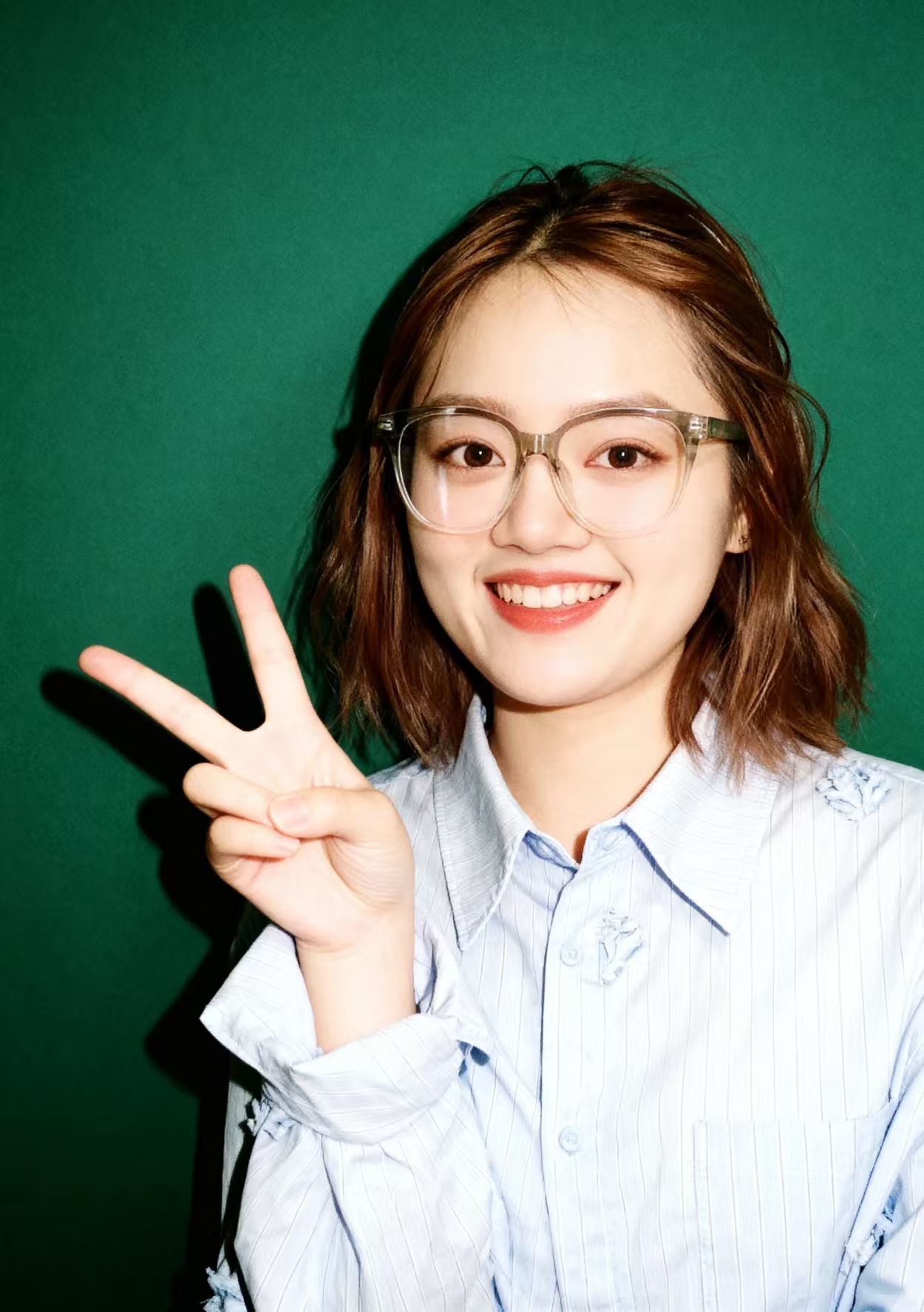 图片展示了一位年轻女性,她面带微笑,右手比出“V”字手势。她戴着眼镜,穿着浅蓝色条纹衬衫,衬衫上有白色花朵图案。背景为纯绿色。这张图片位于文档中“大头贴机器——存量机器焕发新生”案例的生成图对比部分,用于展示经过优化处理后的效果,与上下文提到的“仅凭一张拍完的图片,就可以生成和优化出数十套原本在摄影馆要花大几百才能产出的照片效果”相呼应。